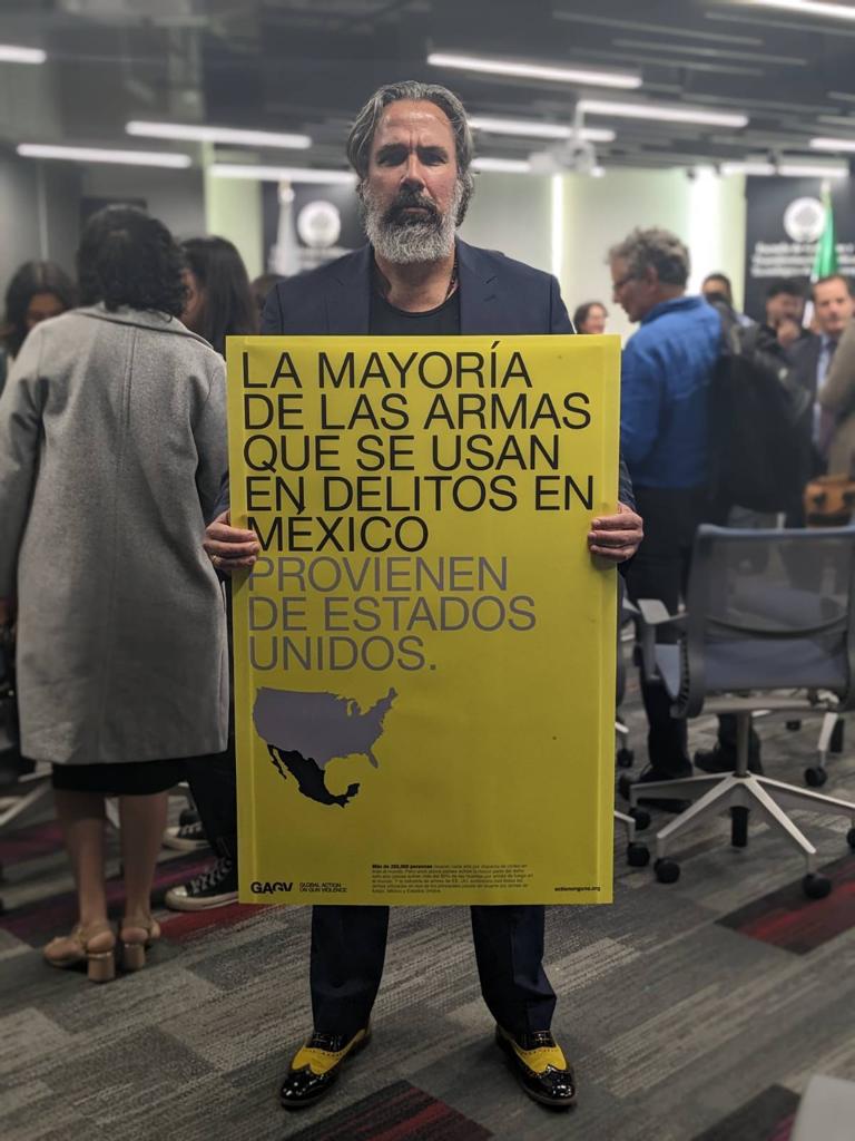 A bearded man stands in a room holding a large yellow and grey poster that says in Spanish, "La mayoria de las armas que se usan en delitos en Mexico provienen de Estados Unidos."