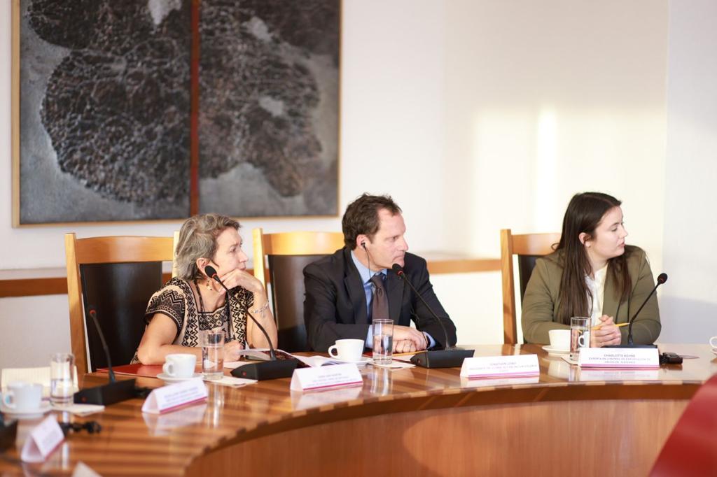 Two women and a man look to their left, listening to a speaker while sitting at a round table in a room. The man sits in the middle. 